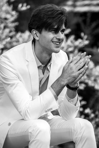 Black-white portrait of young handsome fashionable man in white suit against nature background