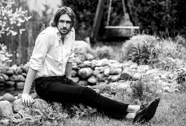 Black-white outdoor portrait of elegant long hair handsome man siting near pond.