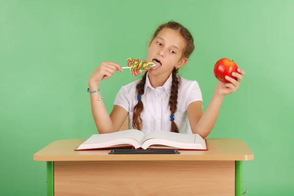 Girl in a school uniform sitting at the desk and choose candy or an apple