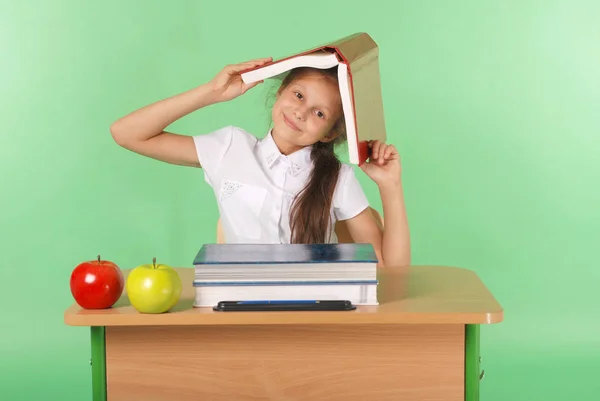 Education, people, children and school concept - young school girl sitting at a desk with a book on her head
