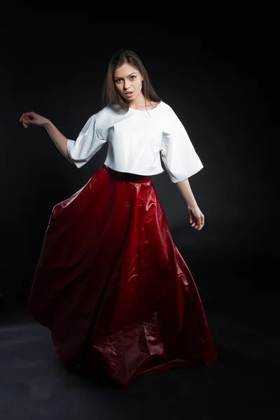 Beautiful young girl in a white blouse and a long skirt red on a black background. Studio portrait.