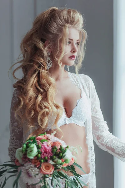 Morning of the bride. Beautiful young woman in white negligee staying near the window with a bouquet
