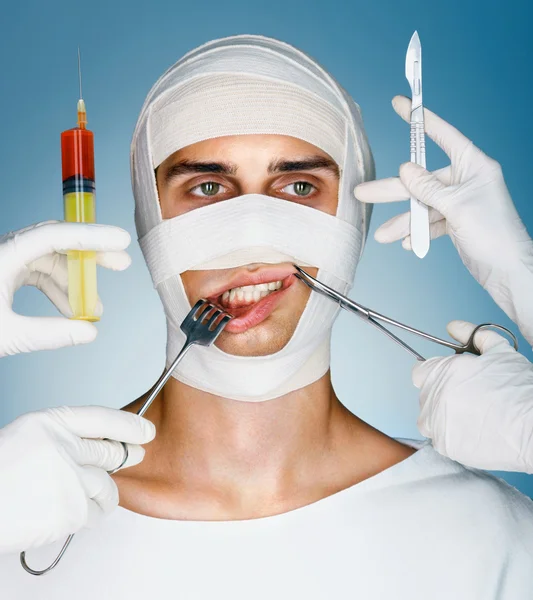 Man wrapped in medical bandages while doctors with syringes, surgical clamp, hook and scalpels near his face.