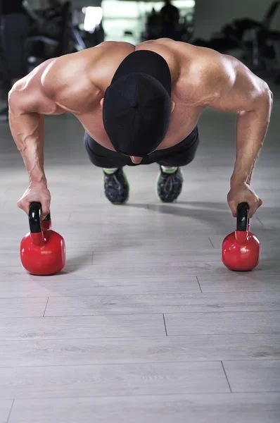 Handsome powerful athletic man performing push ups with kettle bell. Strong bodybuilder with perfect shoulders, biceps and triceps.