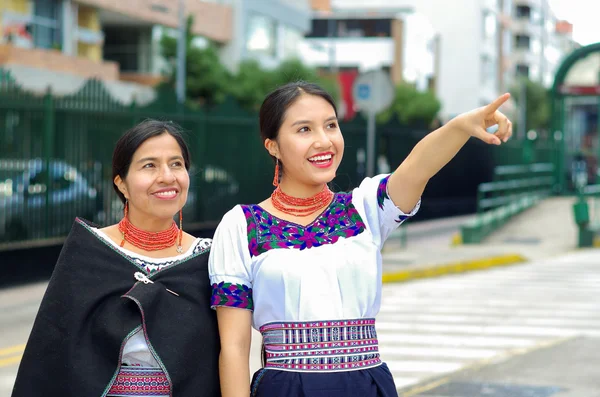 Beautiful hispanic mother and daughter wearing traditional andean clothing, waiting for bus at public station while pointing around, smiling happily, outdoors environment