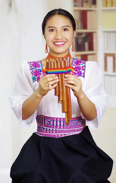 Portrait of young pretty woman wearing beautiful traditional andean clothing, sitting down with while playing the harmonica, bookshelves background