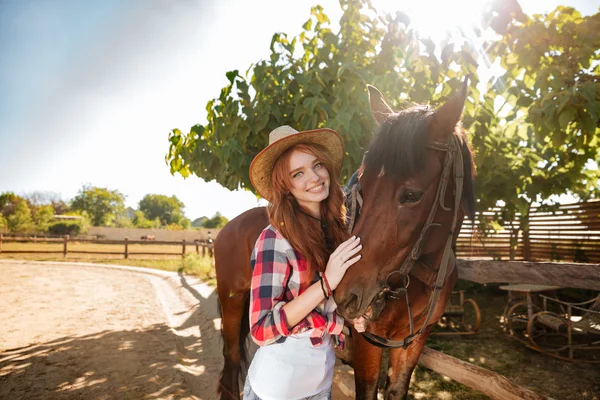 Cheerful young woman cowgirl standing with her horse in village