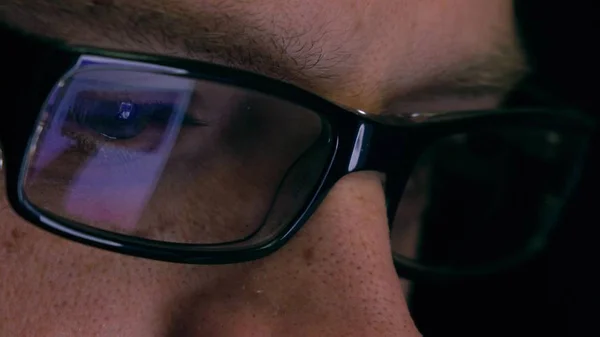 Eyes of serious young man in black rim glasses using his tablet computer. Screen and finger reflecting in the glass