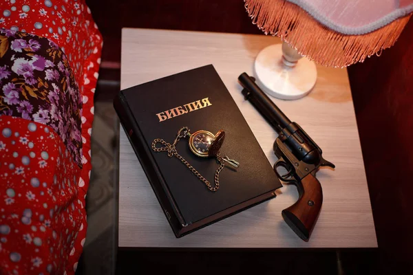 The bible is on the nightstand next to the bed next to the gun with a gold watch
