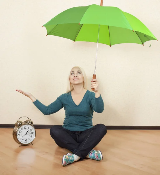 Girl with a green umbrella sits on the floor next to big clock