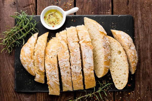 Fresh homemade ciabatta bread sliced on stone slate board, olive oil and rosemary on wooden background