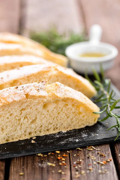 Fresh homemade ciabatta bread sliced on stone slate board, olive oil and rosemary on wooden background