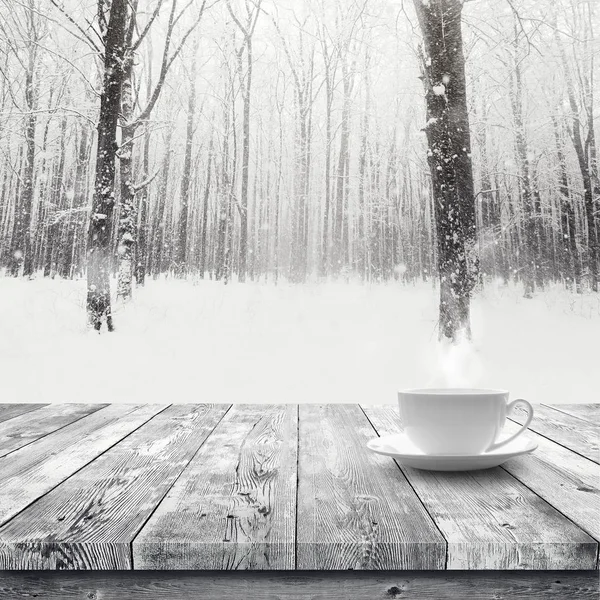 Hot drink in the cup on wooden table over winter snow covered forest. Beauty nature background