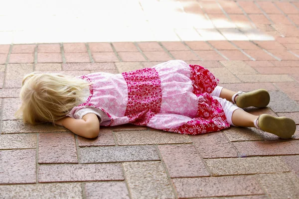 Little blond girl in pink dress lies on park path