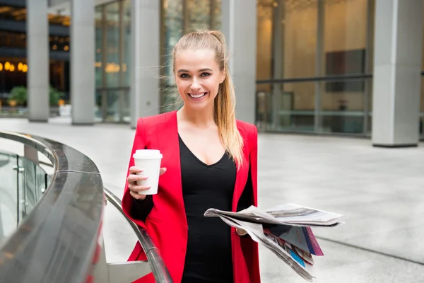 Portrait of cheerful business woman holding coffee and newspaper and enjoying her brake out of office building.