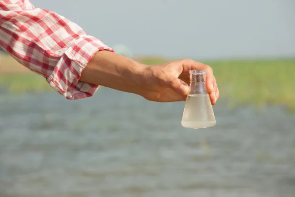 Water Purity Test. Hand holding chemical flask with liquid, lake or river in the background.