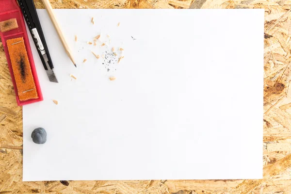 Pencil with kneaded eraser, sandpaper block and cutter knife with shavings, on blank white sheet of paper, on flakeboard