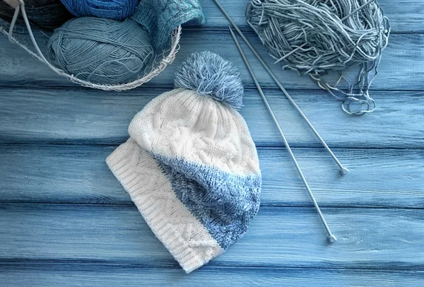 Knitted cap with yarn, needles and metal basket on wooden background