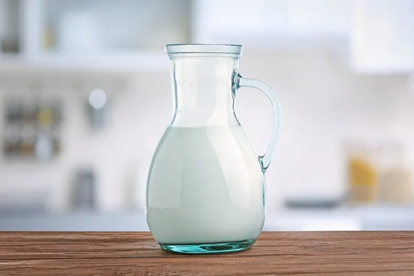 Glass jug of milk on wooden table against blurred kitchen background. Dairy concept.