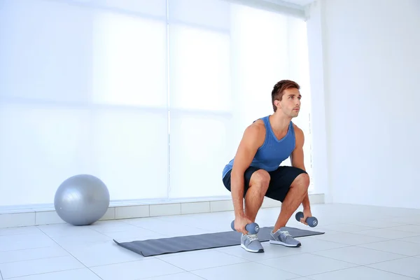 Young man doing exercises with dumb-bells on rug indoor