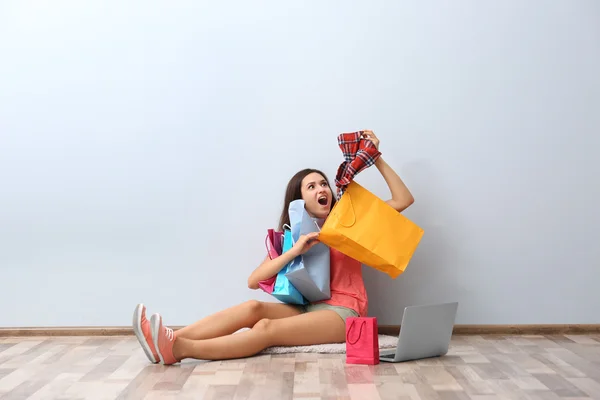 Happy young woman with colorful shopping bags and laptop sitting on floor