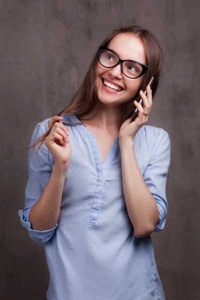 Portrait of smiling woman with glasses speaking by cellphone near grey background wall