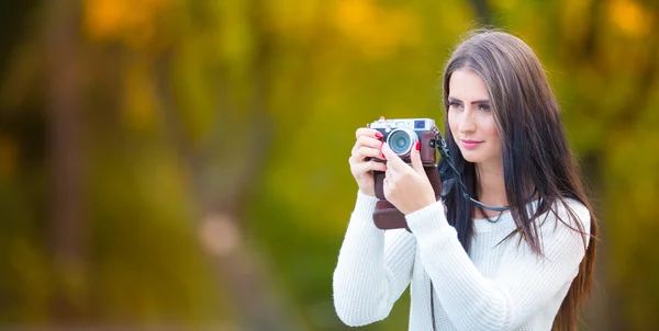 Young attractive brunette woman holding the retro camera in outdoors. Beautiful young girl shooting with retro camera in the autumn sunny day