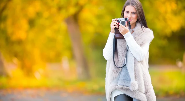 Young attractive brunette woman holding the retro camera in outdoors. Beautiful young girl shooting with retro camera in the autumn sunny day