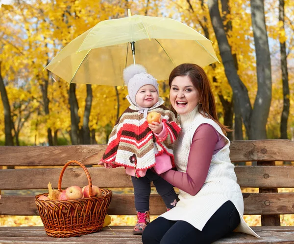 Woman with child girl in autumn city park sit on bench with apples basket and umbrella and having fun, happy family