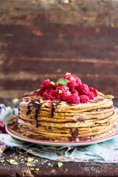 Stack of wheat golden pancakes or pancake cake with freshly picked raspberry, chopped pistachios, chocolate sauce on a dessert plate, selective focus