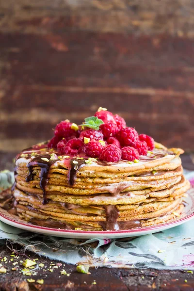 Stack of wheat golden pancakes or pancake cake with freshly picked raspberry, chopped pistachios, chocolate sauce on a dessert plate, selective focus