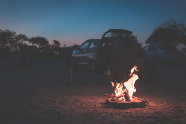 Burning camp fire at dusk in camping site, Botswana, Africa. Summer adventures and exploration in the african National Parks. Selective focus on fire, cars and tents out of focus in the background. To
