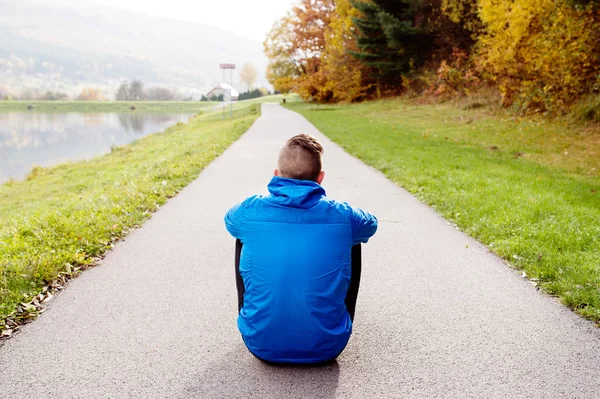 Young runner in park sitting on asphalt path, rear view