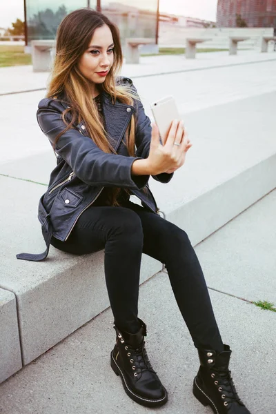 Beautiful smiley young woman with nice make-up is sitting on the concrete steps and doing live snap-chat vlog video on her smart-phone. Copy-space area for your text or advertisement text