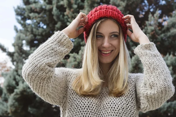 Blonde woman placing her red knitted wool cap, against of fir tr