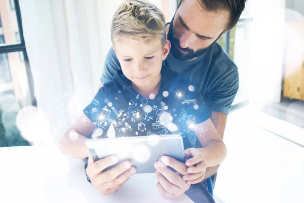 Father and his little son playing together on mobile computer, resting indoor.Bearded man with young boy using tablet PC in sunny house.Childhood dreams icons concept.Horizontal, blurred background.