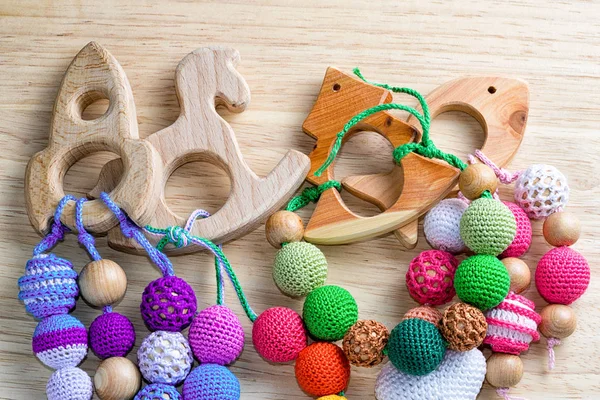 Four necklaces made from knitted beads and wooden toys for the baby sitting in a sling. Toys on a wooden table.