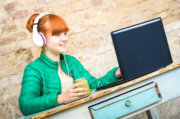 Hipster redhead woman with headphone using laptop drinking cocktail and having fun at work - Modern concept of professional engagement connected with happy and productive attitude - Focus on girl face