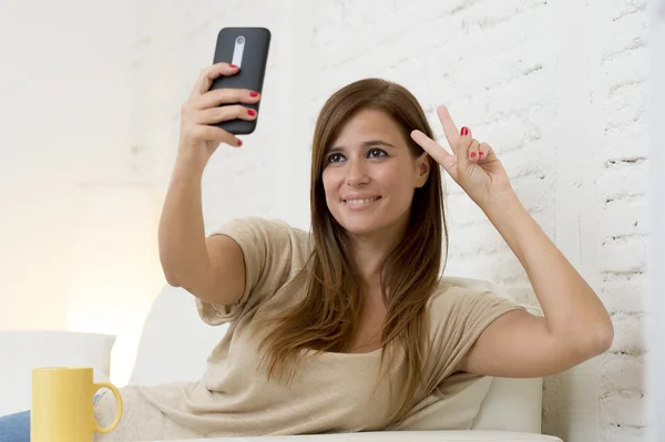 Attractive 30 years old woman playing on home sofa couch taking selfie portrait with mobile phone