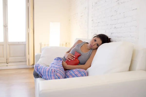 Young beautiful hispanic woman holding hot water bottle against belly suffering menstrual period pain