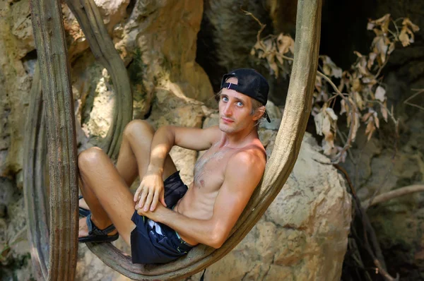 Shirtless young man relaxing and leaning against tree or a liana in front of cave