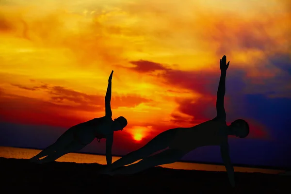 Yoga people training and meditating in warrior pose outside by beach at sunrise or sunset.