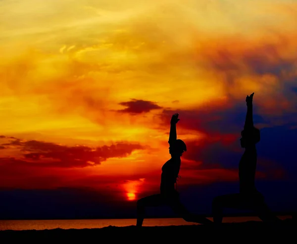 Yoga people training and meditating in warrior pose outside by beach at sunrise or sunset.