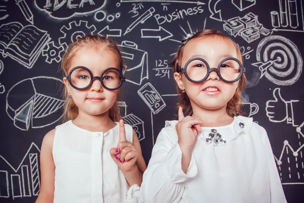 Two little smart girls in glasses lifting finger up on a background of wall with business or school picture