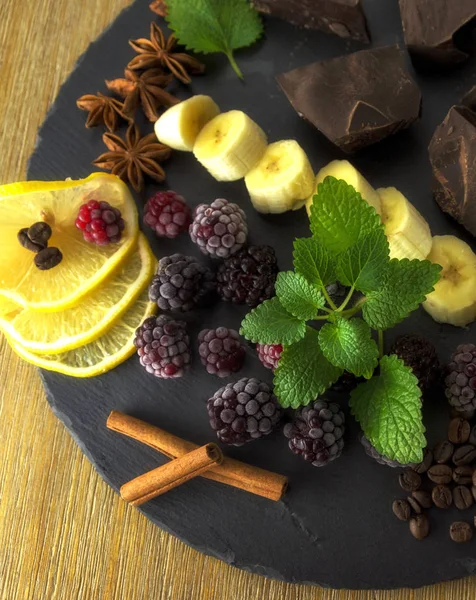 Exposition close up of fresh organic fruit and chocolate, half of lemon, sliced banana, mint leaf, cranberry, blackberry and coffee grape on black background,  rock board, wooden table, healthy food.
