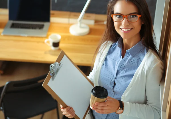 Young woman standing near desk with laptop holding folder and cup of coffee