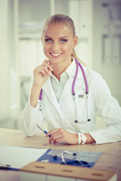 Beautiful young smiling female doctor sitting at the desk and writing.