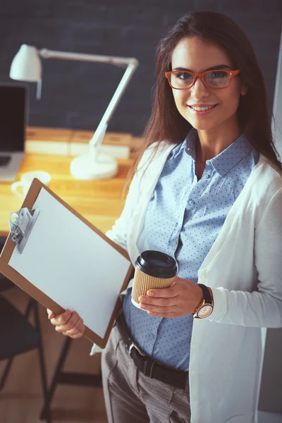 Young woman standing near desk with laptop holding folder and cup of coffee