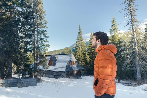 Handsome young man looking at astonishing view in valley with mountain hut and conifer forest