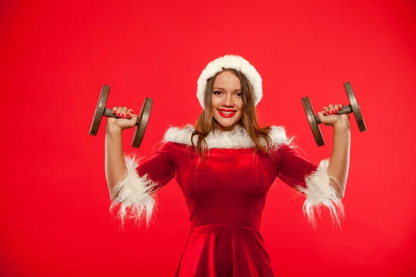Christmas, x-mas, winter, happiness concept - Bodybuilding. Strong fit woman exercising with dumbbells in santa helper hat , over red background. Muscular blonde girl lifting weights studio shot